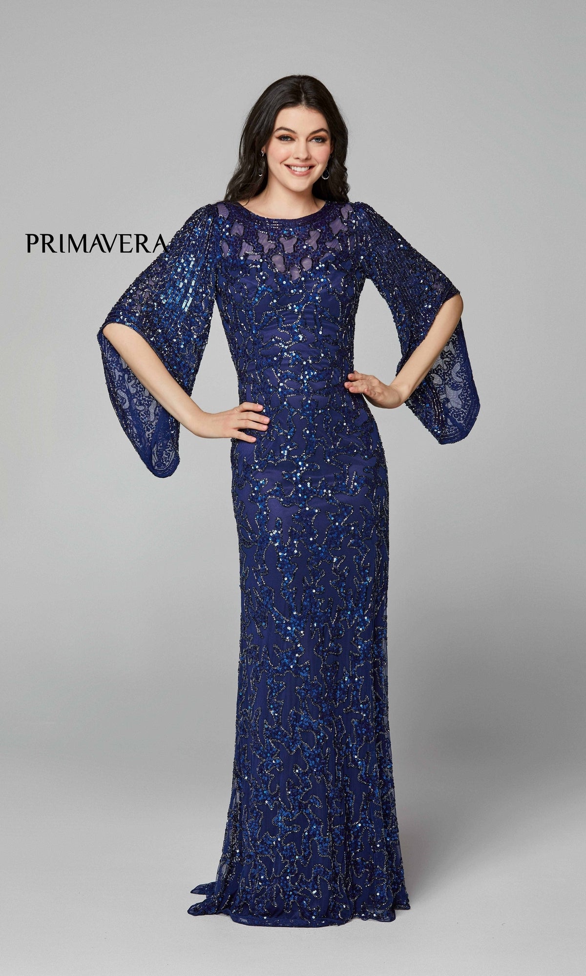 Woman in the Long Bell Sleeve Beaded Evening Gown 9713, a navy floor-length dress with beading and sheer bell sleeves, stands against a gray background.